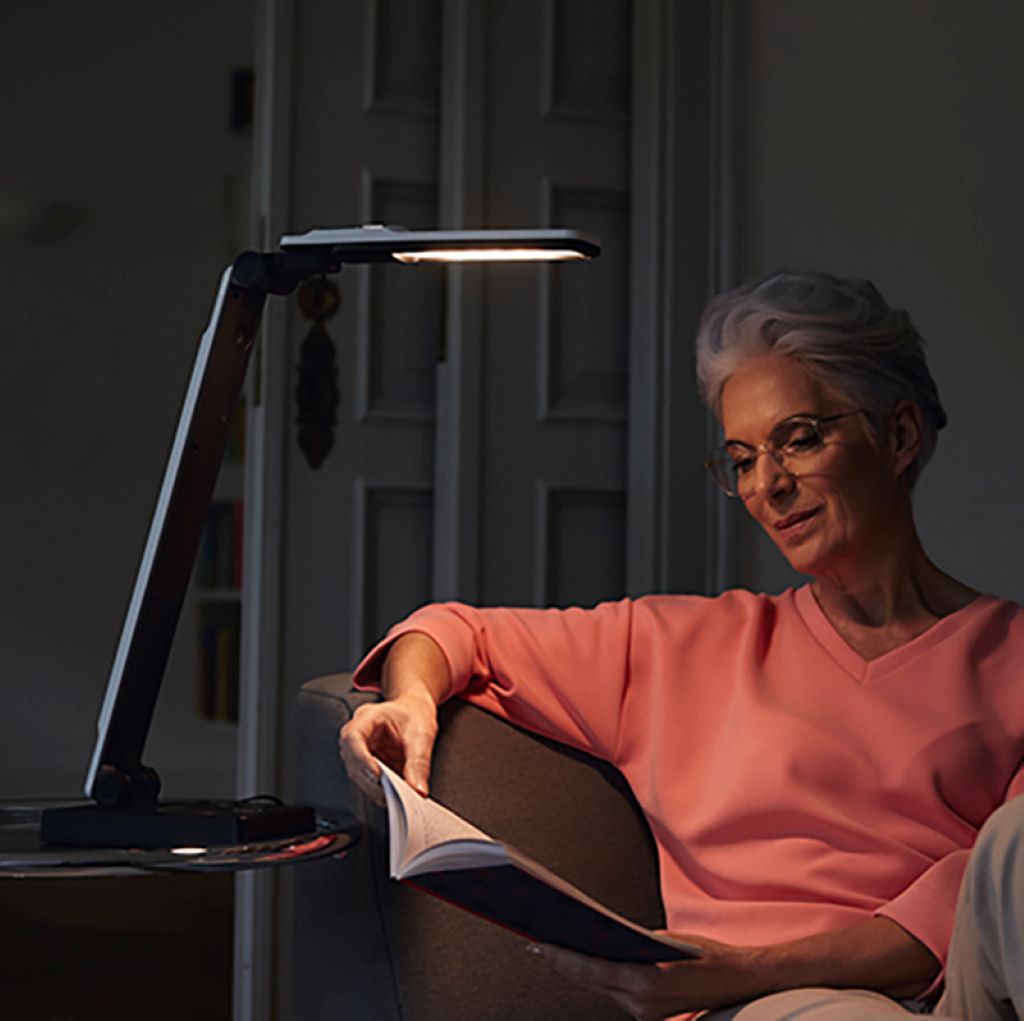 Image of a woman sitting on a couch in a dark room with the Elumentis LED Lamp beside her on a side table, turned on with the light right on her book that she is reading.