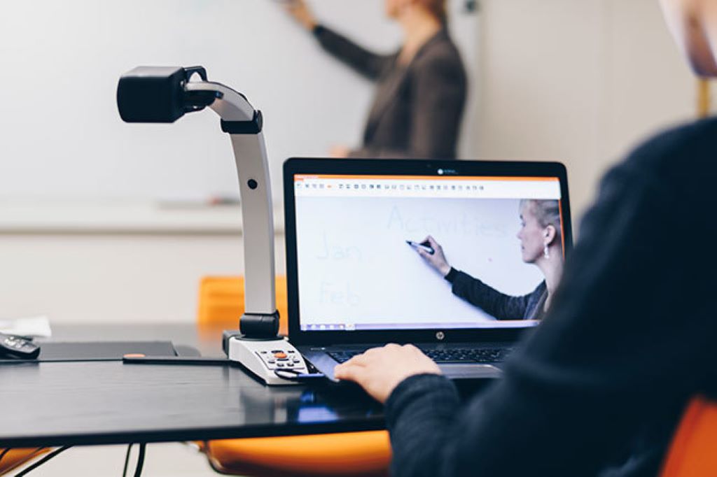 Image of a student using the MagniLink S Premium 2 on their desk in a classroom to view a magnified view of the teacher and what she is writing on the whiteboard. 