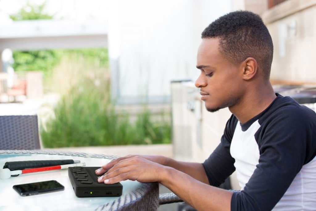 image of a student using the Brailliant BI 20X while sitting at a table that is outside, with their cane and cell phone besides the braille display on the table.