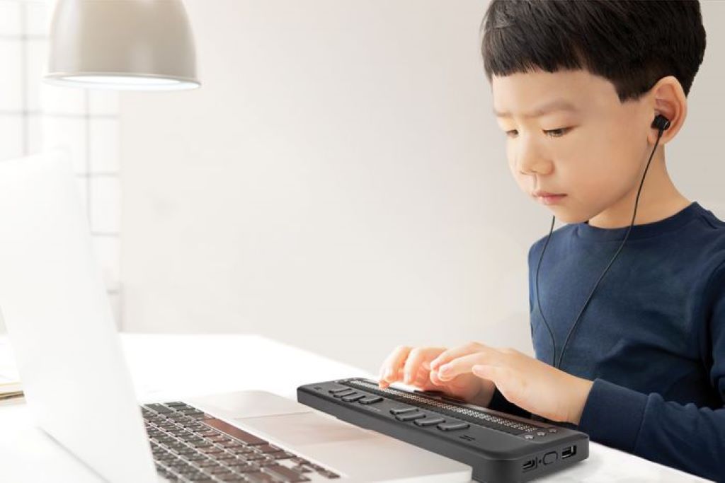 image of a young student with headphones in, sitting at a table, typing on the brailliant bi 40x braille display