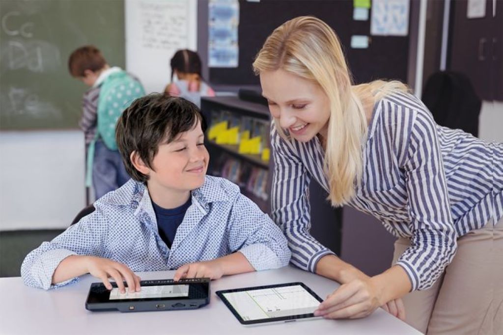 image of a student at his desk using the braillenote touch plus with his teacher standing beside him while viewing an equation on a tablet.