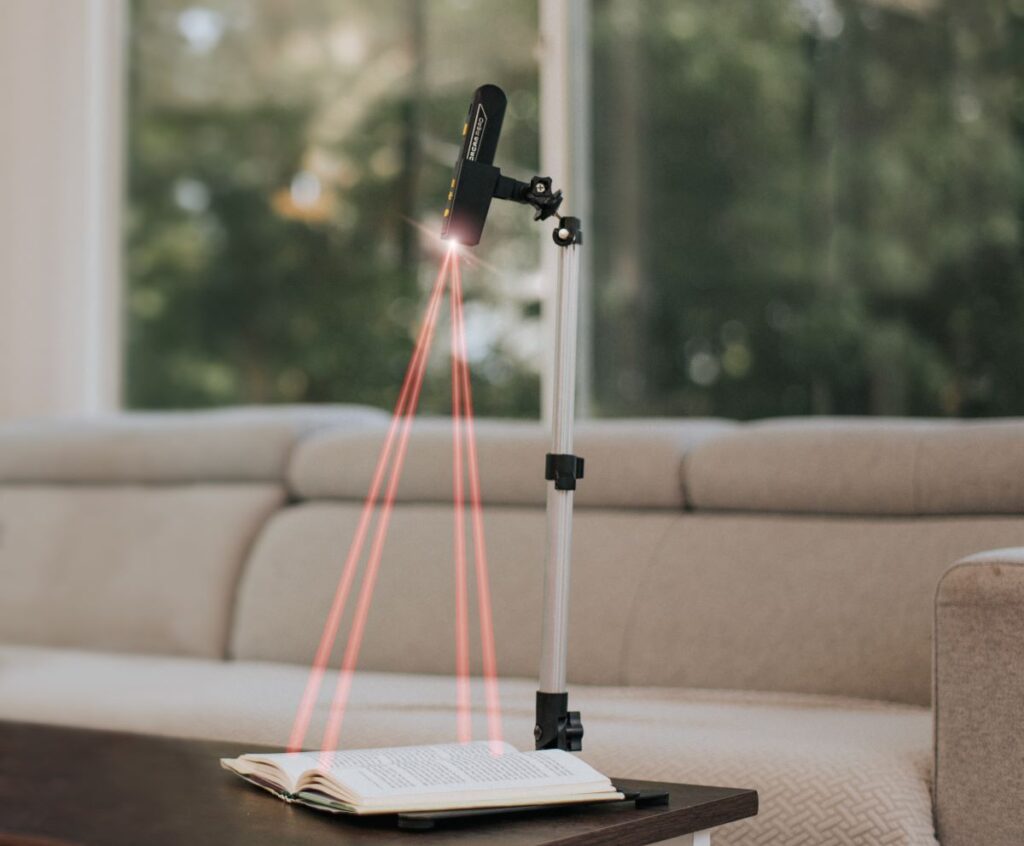 image of the orcam read 3 in its stand sitting on someones coffee table in a living room scanning the text on a book underneath