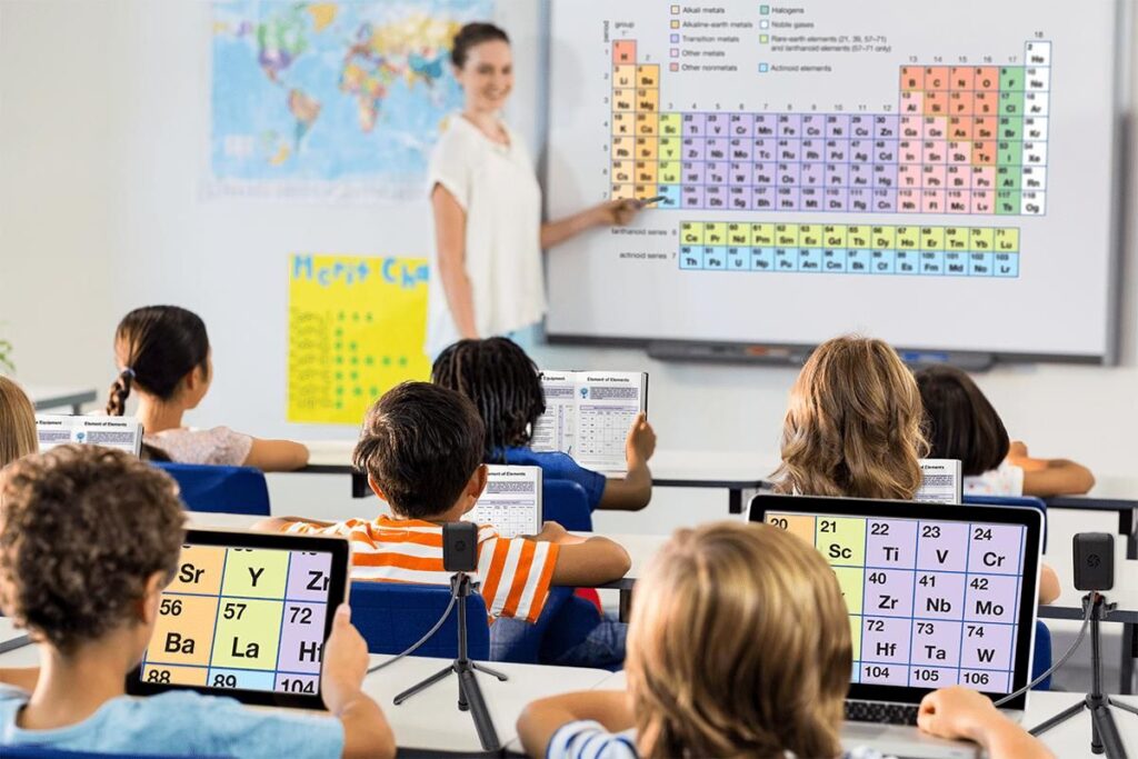 image of students in a classroom sitting at desks while the teacher is at the front showing the periodic table and two students are using coast camera's to view a magnified picture of the periodic table on their iPads